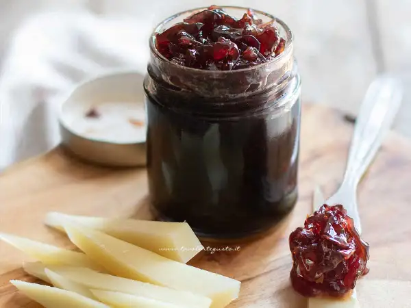Un vasetto di marmellata di cipolle rosse, con un cucchiaino che ne assaggia un po' su una fetta di formaggio.
