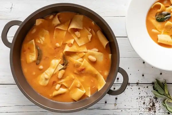 Un saporito piatto di maltagliati con fagioli, servito in una padella scura e una ciotola bianca, con erbe aromatiche e pepe nero a guarnire.