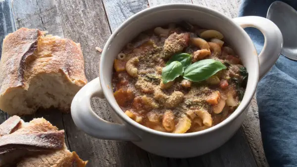 Una ciotola di pasta e fagioli in scatola, guarnita con basilico fresco e parmigiano, accanto a pane rustico su un tavolo di legno.