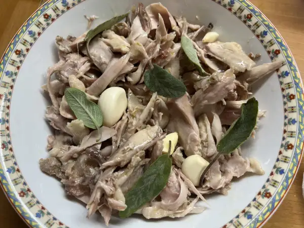 Piatto di tonno di coniglio sfilacciato, condito con aglio e foglie di salvia fresca, servito in una ciotola decorata.