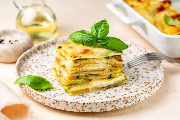 Una fetta di parmigiana di zucchine bianca alla napoletana, stratificata e dorata, guarnita con basilico fresco.