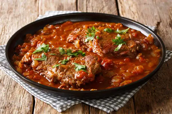 Carne alla pizzaiola morbida, succulenta e profumata, servita in un piatto scuro su un tavolo di legno.