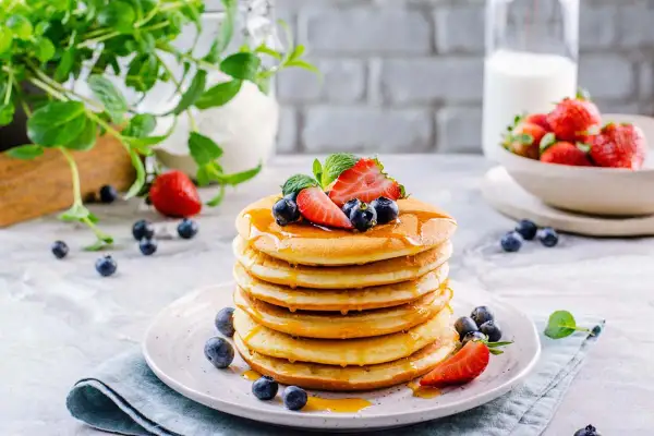 Pila di soffici pancake senza glutine e lattosio, guarniti con fragole, mirtilli e sciroppo d'acero.