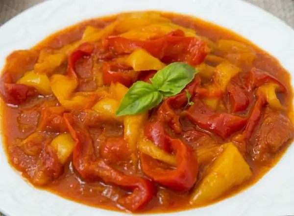 Peperonata ricetta della nonna: peperoni rossi e gialli cotti lentamente in un sugo saporito, guarniti con una fogliolina di basilico fresco.