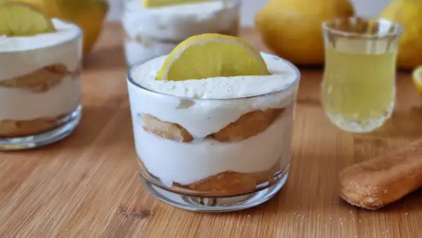 Delizioso tiramisù al limoncello in bicchieri, guarnito con una fetta di limone. Accanto, un bicchierino di limoncello e savoiardi.