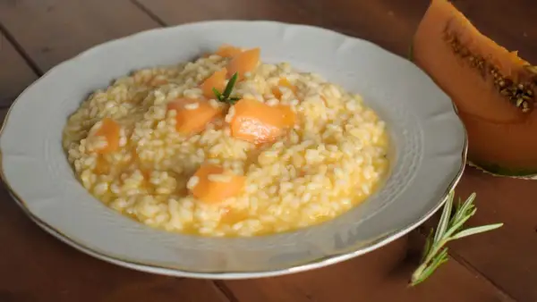 Un cremoso risotto con pezzi di melone e un rametto di rosmarino, accanto a una fetta di melone.
